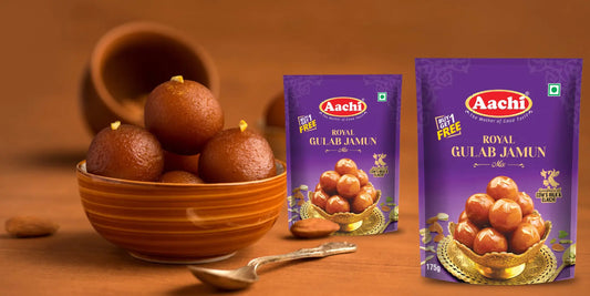 Aachi Royal Gulab Jamun Mix packs showcased next to a bowl of soft, golden-brown gulab jamuns topped with pistachios, elegantly set on a wooden surface with a spoon beside it.